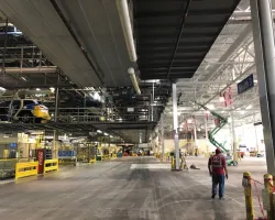 Large industrial warehouse with assembly lines, overhead conveyor systems, and a worker walking inside.