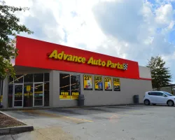 Advance Auto Parts store exterior with red signage, promotional posters, parked cars, and partly cloudy sky.