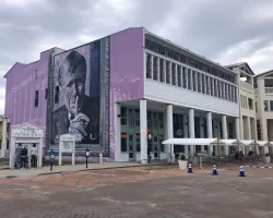 Large purple building with a mural of an elderly man shushing and a tourist information booth outside under cloudy sky.