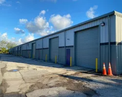 Long metal warehouse building with multiple closed roller doors and yellow bollards under a blue sky.