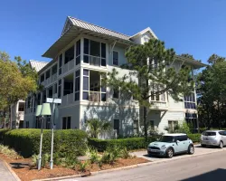 Modern three-story residential building with balconies surrounded by trees and parked cars on a sunny day
