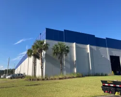 Modern industrial building with blue accents, palm trees, green lawn, and trailer parked outside under clear sky