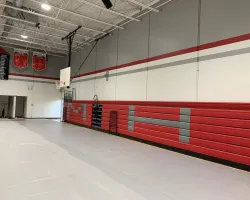 Empty gymnasium with red and gray retractable bleachers and basketball hoop on a polished floor.