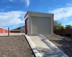 Self-storage unit with a closed roll-up door at the top of a concrete ramp under a blue sky with clouds.