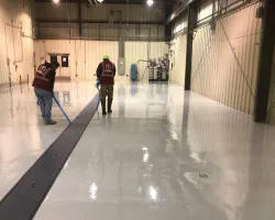 Two workers applying epoxy coating to a clean industrial warehouse floor using rollers under bright lights