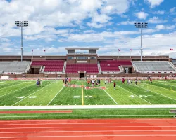 Wide view of a football field with players practicing and empty stadium seating under a partly cloudy sky