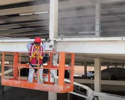 Construction worker in red helmet using a lift to clean or repair a white concrete parking structure beam.
