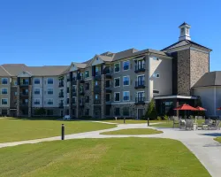 Large modern apartment complex with green lawn, walking paths, and outdoor seating with red umbrellas under clear sky