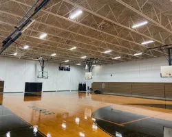 Empty indoor basketball court with wooden floor, multiple hoops, bright ceiling lights, and bleachers on side.
