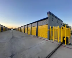 Wide view of a storage facility with yellow roll-up doors and gated entrance under a clear sky at sunset.