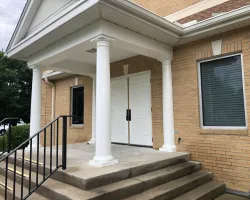 Entrance to a brick building with white double doors, four white columns, and concrete steps with black railing.