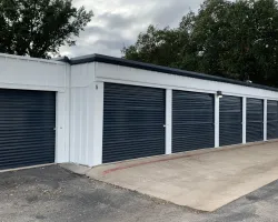 Row of white storage units with dark blue roll-up doors under a cloudy sky and surrounded by trees.