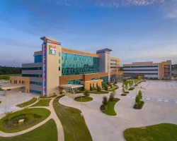 Modern hospital building with glass facade, colorful pillars, surrounding greenery, and spacious parking lot under blue sky.