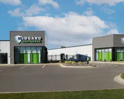 Modern Midgard Self Storage facility with green doors, clear sky, and ample parking spaces in front.