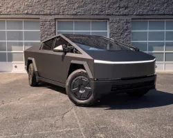 Matte black Tesla Cybertruck parked outside a building with three white garage doors on a sunny day.