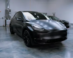 Matte black Tesla Model Y parked in a spacious indoor showroom with polished concrete floors and other cars around.