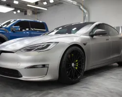 Silver Tesla Model S with black wheels parked inside a modern garage with a blue truck in the background