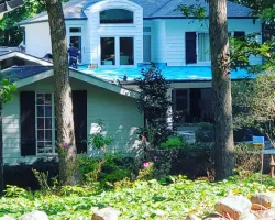 Two-story white house with blue roof under construction surrounded by trees and greenery.