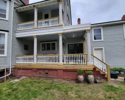 Front entrance of a beige two-story house with white porch columns, brick steps, black shutters, and green lawn.