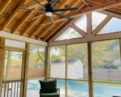 Cozy screened porch with wooden ceiling, black ceiling fan, green chairs, coffee table, and pool cover outside.