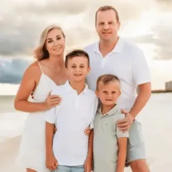 Family of four posing on a sandy beach with cloudy sky in the background, wearing casual summer clothes.