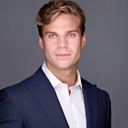 Professional headshot of a young man in a navy suit and white shirt with a neutral gray background