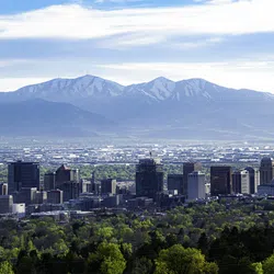 Panoramic city skyline surrounded by lush greenery with towering mountains in the background under a bright blue sky