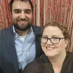Man in suit and woman with glasses smiling in front of pink and gold shimmering fringe backdrop