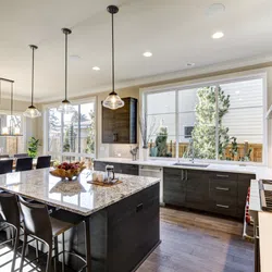 Modern kitchen with dark cabinets, large granite island, pendant lights, and dining area with black chairs and large windows.