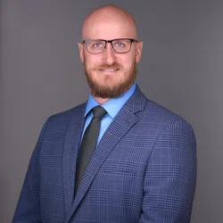Smiling bald man with glasses and beard wearing a blue plaid suit, light blue shirt, and black tie on gray background