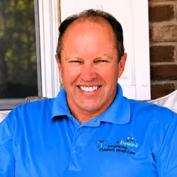 Smiling man in bright blue polo with Children’s Health Care logo sitting on porch with brick wall background