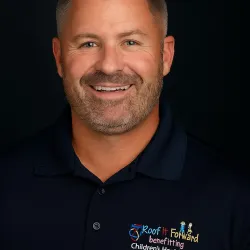 Smiling man with short gray hair and beard wearing a black polo with Roof It Forward Children’s Health Care logo.