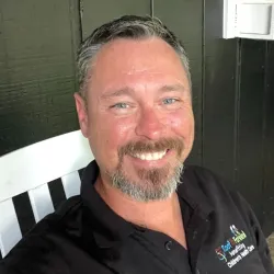 Smiling man with gray beard wearing black collared shirt sitting on white chair against black wood panel wall.