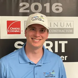 Smiling man in blue shirt and white hat with Children’s Health Care logos in front of a 2016 Owens Corning award banner.