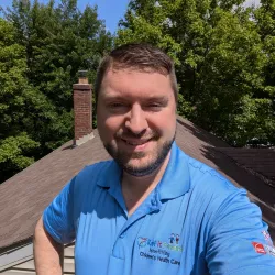 Smiling man in blue polo shirt with charity logo stands outdoors on a rooftop surrounded by green trees
