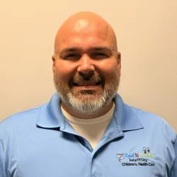 Smiling bald man with gray beard wearing light blue polo shirt with Roof It Forward Children's Health Care logo