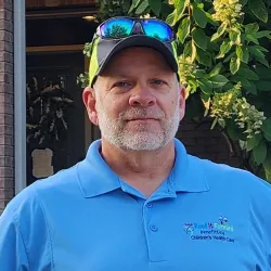 Man wearing blue polo with charity logo and neon cap with reflective sunglasses outdoors near greenery and brick wall.