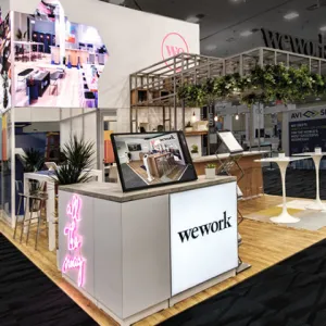 Modern WeWork exhibition booth with digital displays, neon signage, plants, and wooden flooring at a trade show.