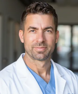 Confident male doctor in white coat and blue scrubs smiling in a bright medical facility hallway..
