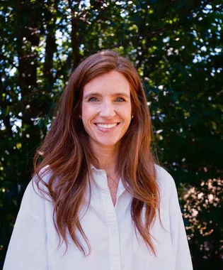 Smiling woman with long red hair wearing a white shirt stands outdoors with green foliage background..