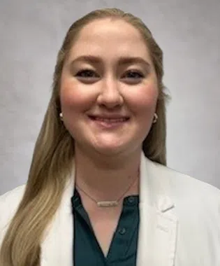 Smiling woman with long blonde hair wearing a white coat and dark blouse against a neutral background.