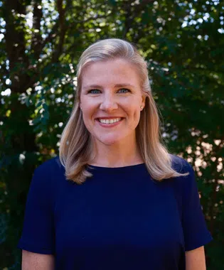 Smiling blonde woman in navy blue shirt standing outdoors with green leafy trees in the background.