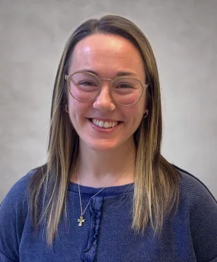 Smiling woman with glasses, nose ring, cross necklace, and long hair wearing a blue sweater against a plain background..