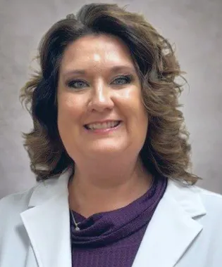 Smiling middle-aged woman with curly brown hair wearing a white coat and purple top against neutral background.
