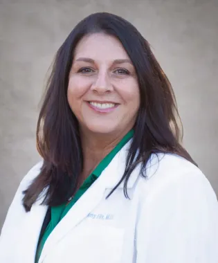 Smiling female doctor with long dark hair wearing a white coat and green shirt on a neutral background.