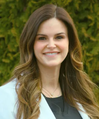 Smiling woman with long wavy hair wearing a white coat standing outdoors with green foliage background.