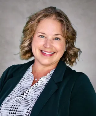 Smiling woman with curly blonde hair wearing a black blazer and patterned white blouse against gray background.