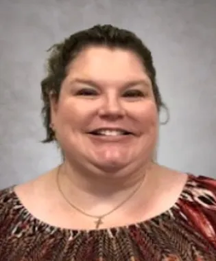 Smiling woman with curly hair wearing a patterned top and a cross necklace against a gray background.