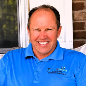 Smiling man in bright blue polo with Children’s Health Care logo sitting on porch with brick wall background