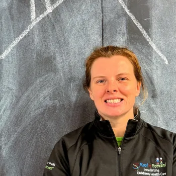 Smiling woman in black jacket stands in front of chalkboard with house roof drawing above her head.